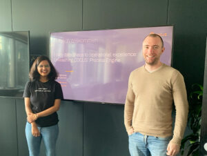 Two people smile at the camera and stand in front of the monitor displaying the presentation.