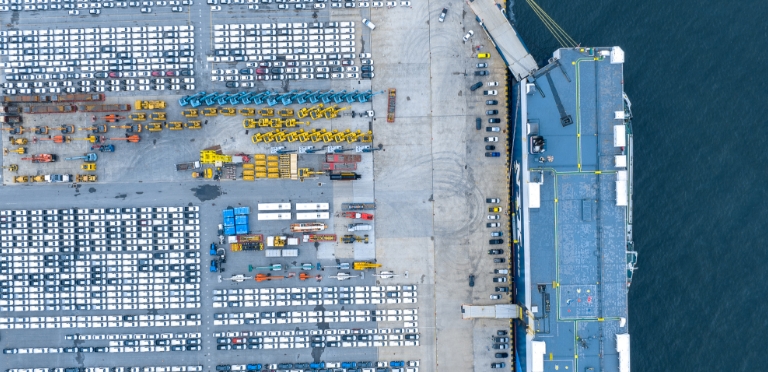 Luftaufnahme eines Hafens mit Reihen von Fahrzeugen und Kränen, die auf eine geschäftige Industrie- oder Logistikumgebung hindeuten.